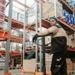 Warehouse worker operating a pallet jack among stacked boxes and metal shelves in an industrial setting.